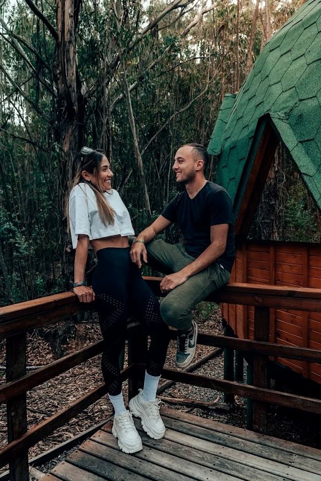 Pareja riendo sentada en la baranda de la terraza de un domo geodésico, rodeada de bosque, en Pesca, Boyacá